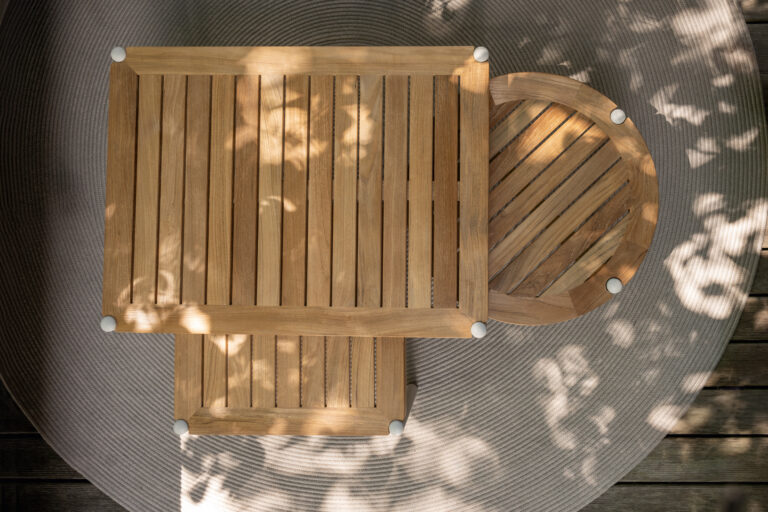 Drei hölzerne Tische – zwei quadratische und ein runder – stehen versetzt zueinander auf einem runden, strukturierten Teppich im Freien. Sonnenlicht und Schatten von Blättern fallen malerisch auf die Szene und erzeugen ein natürliches, ruhiges Ambiente.