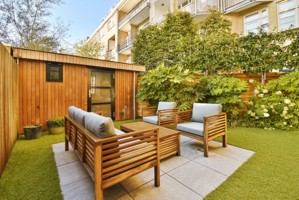 Ein Gartenbereich mit Holzmöbeln und grünem Gras auf dem Boden vor einem Wohnhaus, das von Bäumen umgeben ist.