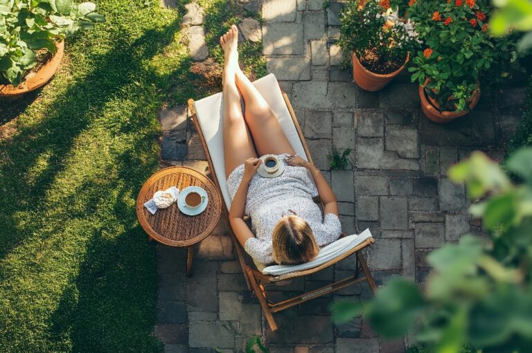 Eine Frau entspannt sich in einem Liegestuhl im Garten und genießt einen Tee an einem sonnigen Nachmittag, umgeben von blühenden Blumen und Grünpflanzen