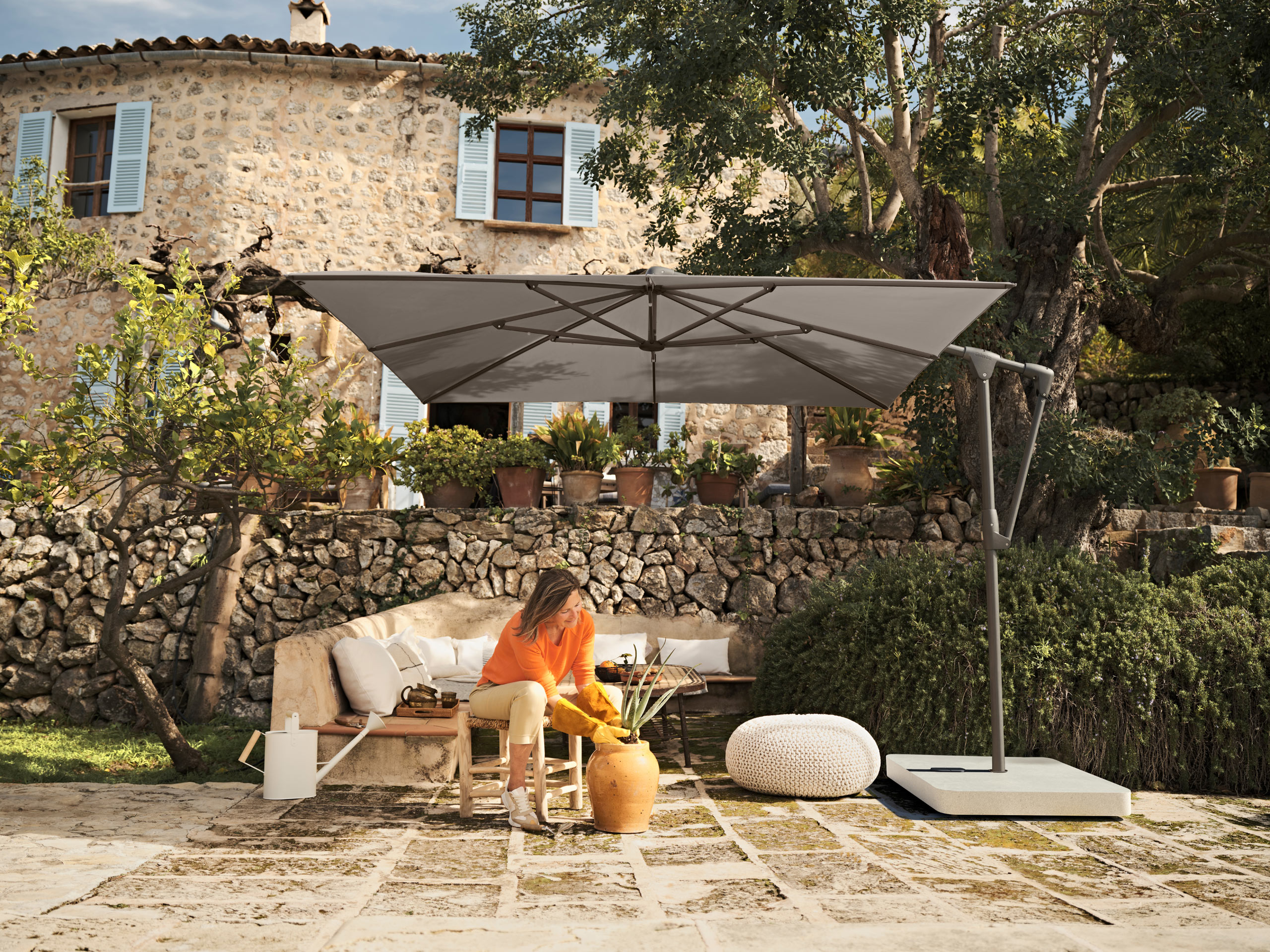 Eine Frau mit blondem Haar sitzt entspannt auf einem Sofa im Garten vor einem Natursteinhaus