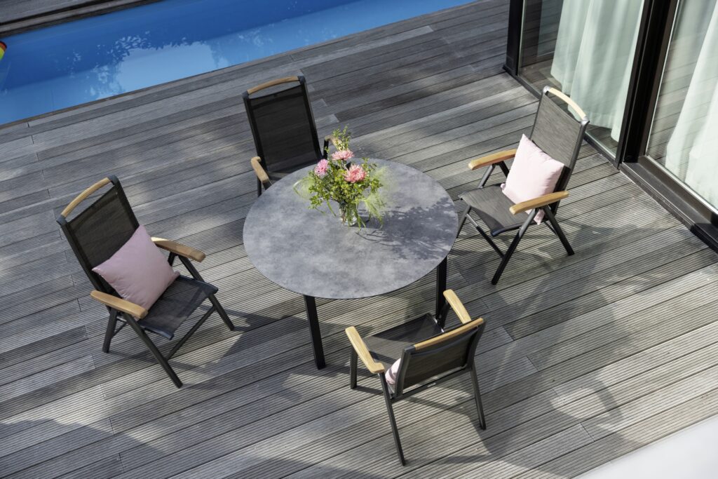 Ein runder grauer Tisch mit Blumenstrauss steht auf einer Holzterrasse neben einem Pool