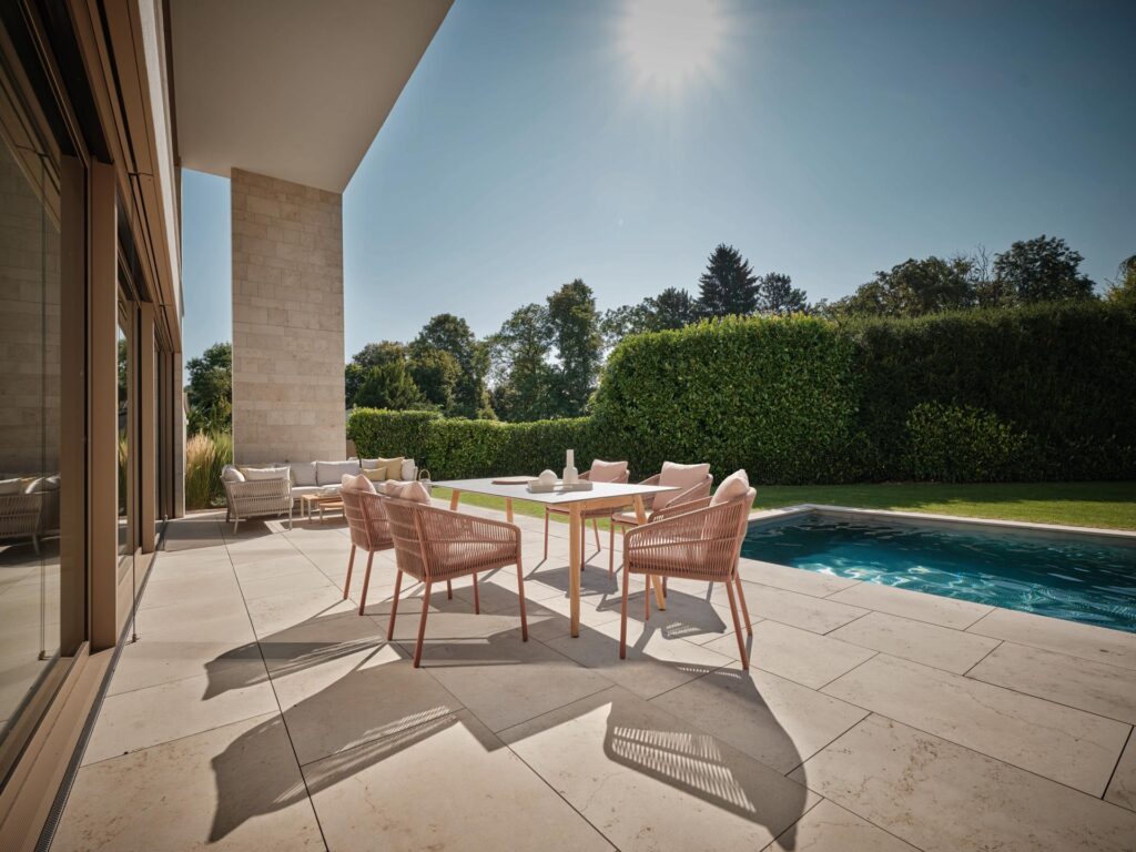 Moderne Terrasse mit beigen Möbeln am Pool und Sonnenschein vor grünen Hecken